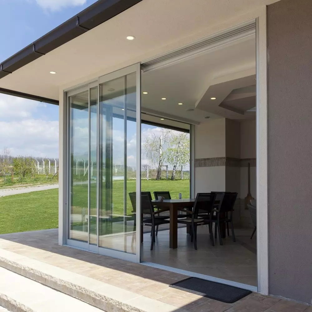 Veranda moderna con tavolo e sedie all’aperto, grandi vetrate scorrevoli e vista su giardino verde soleggiato.