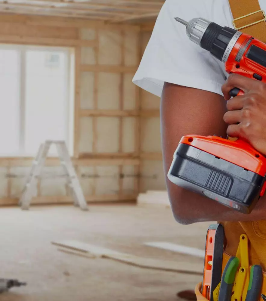 Operaio in cantiere intento alla ristrutturazione di una casa impugna un trapano elettrico arancione, sfondo di pareti in costruzione e scala in legno.